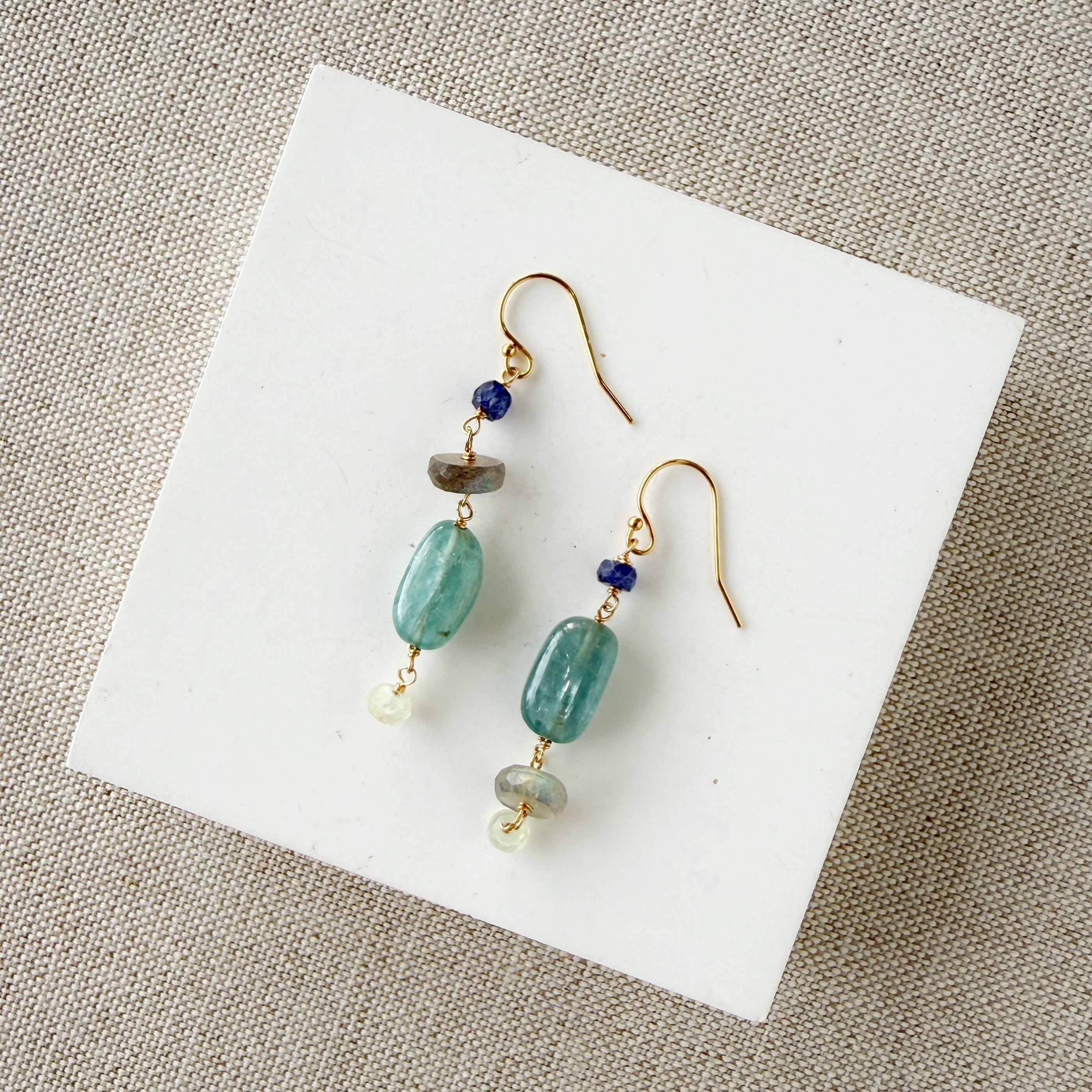 Pair of earrings with blue and teal gemstones on a white card against a beige background
