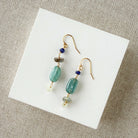 Pair of earrings with blue and teal gemstones on a white card against a beige background