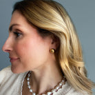 Close-up of a woman wearing gold dome earrings and a pearl necklace against a gray background
