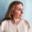 Woman wearing a beige sweater, gold dome earrings and pearl necklace against a light blue background