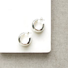 A pair of polished silver crescent-shaped hoop earrings displayed on a white square card over a textured gray fabric background.