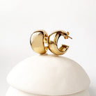 A pair of chunky gold hoop earrings with a polished, rounded organic design, displayed on a stacked white ceramic stand against a clean white background.