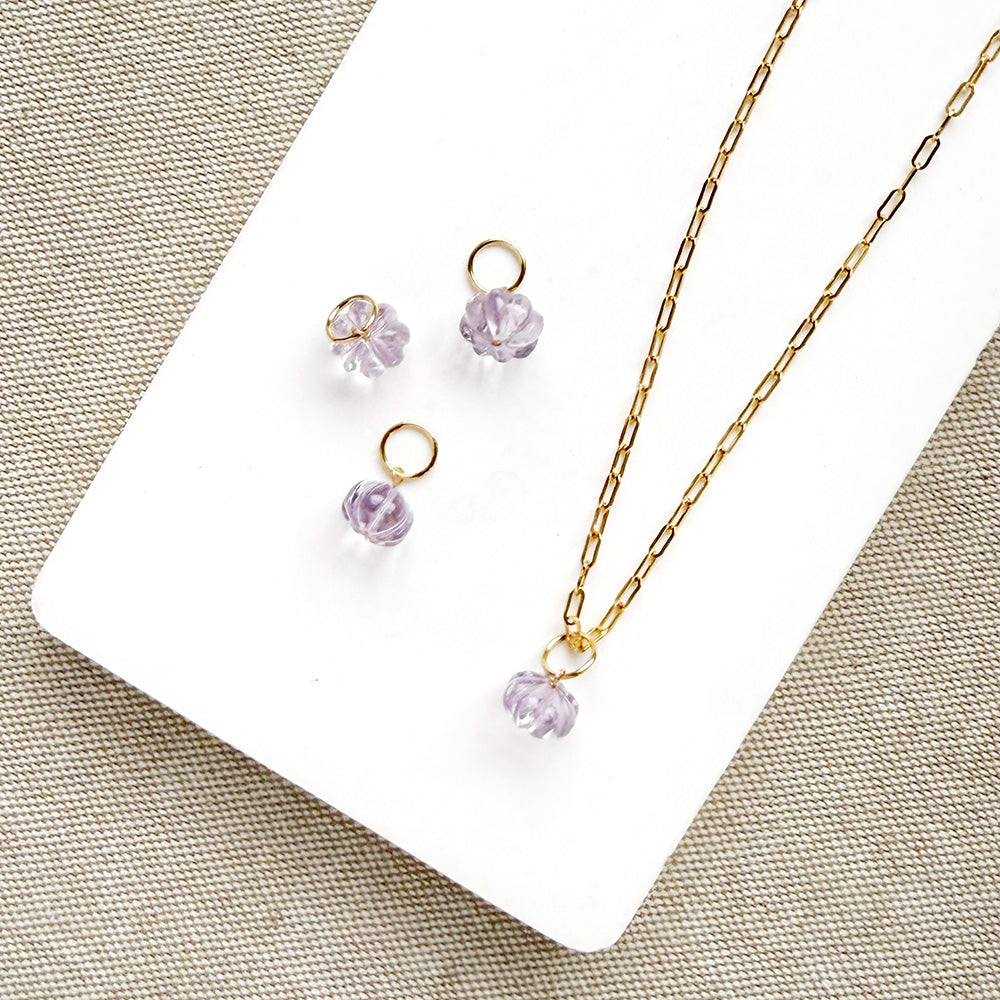 Gold chain necklace and matching drop earrings with small carved amethyst charms, arranged on a white card over a beige textured fabric background.