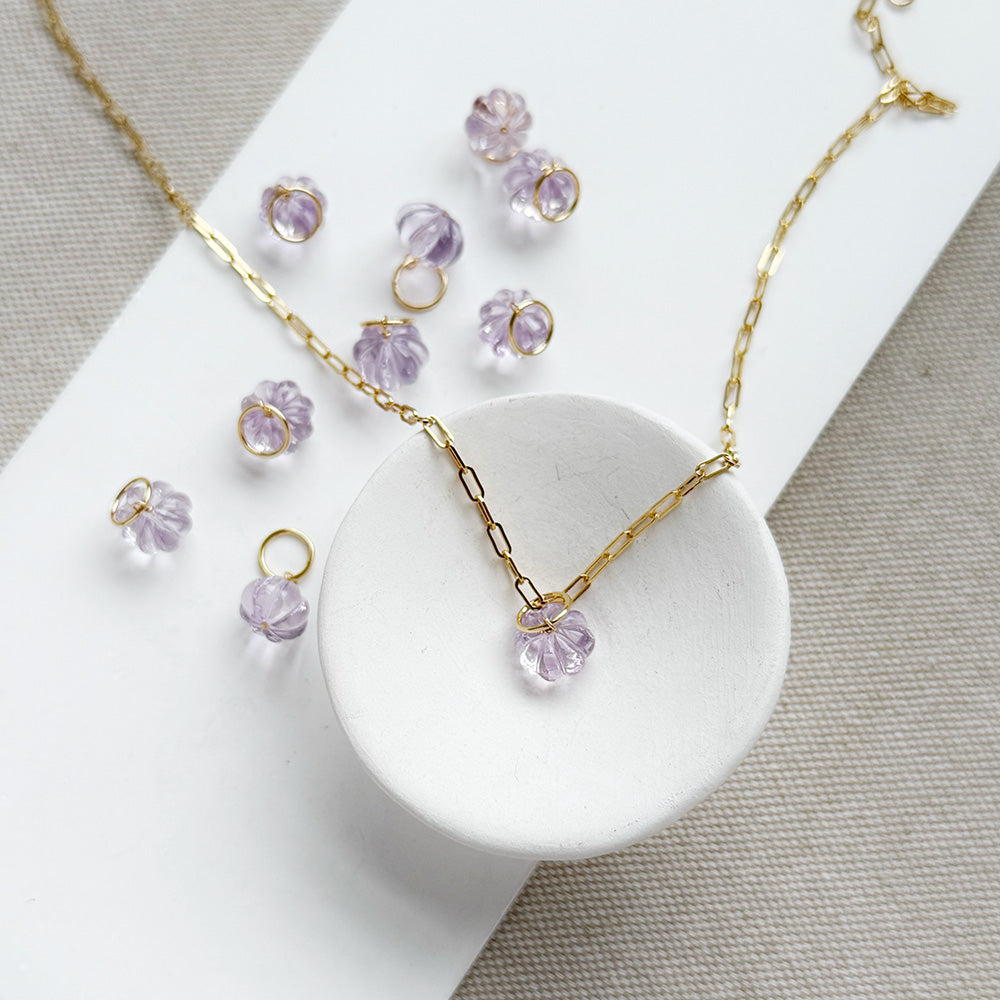 Gold chain necklace and matching drop earrings with small carved amethyst charms, arranged on a white card over a beige textured fabric background.