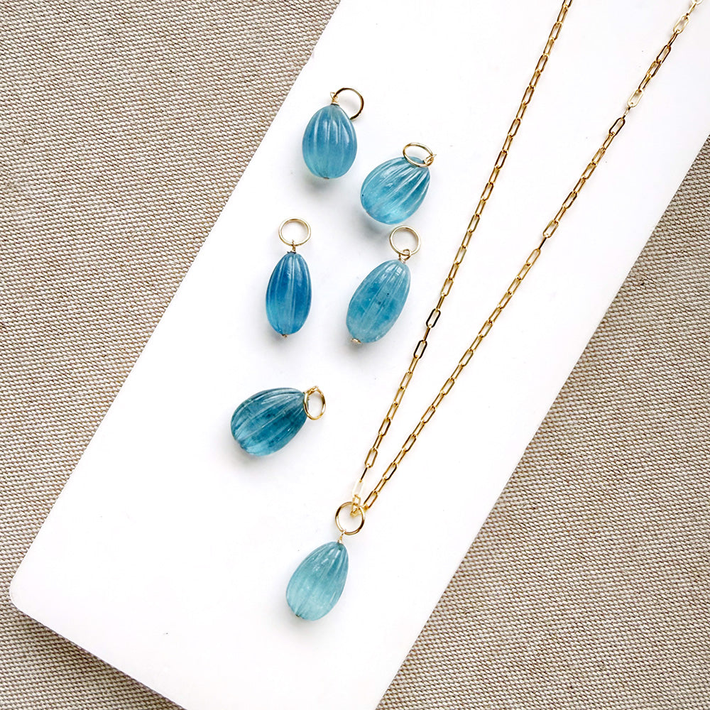 Gold-tone jewelry featuring a teardrop-shaped light blue carved aquamarine charms arranged on a white display block and textured beige fabric.