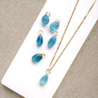 Gold-tone jewelry featuring a teardrop-shaped light blue carved aquamarine charms arranged on a white display block and textured beige fabric.