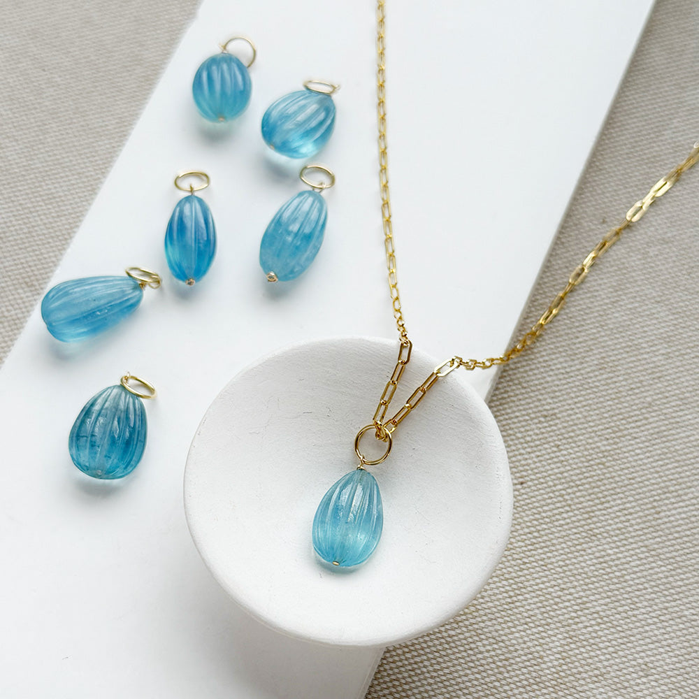Gold-tone jewelry featuring a teardrop-shaped light blue carved aquamarine charms arranged on a white display block and textured beige fabric.