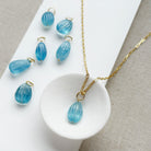 Gold-tone jewelry featuring a teardrop-shaped light blue carved aquamarine charms arranged on a white display block and textured beige fabric.