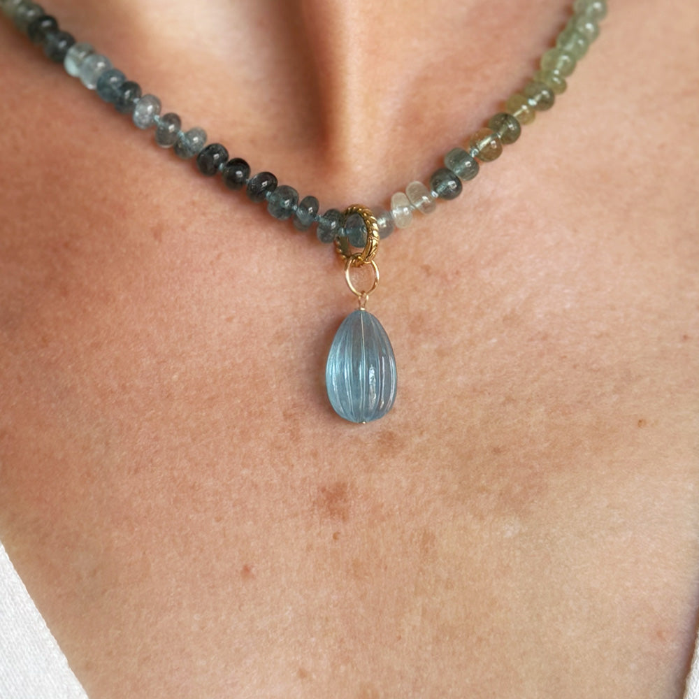 A close-up of a beaded necklace worn on a person's neck, featuring a faceted light blue carved aquamarine charm with gold-tone metal accents.