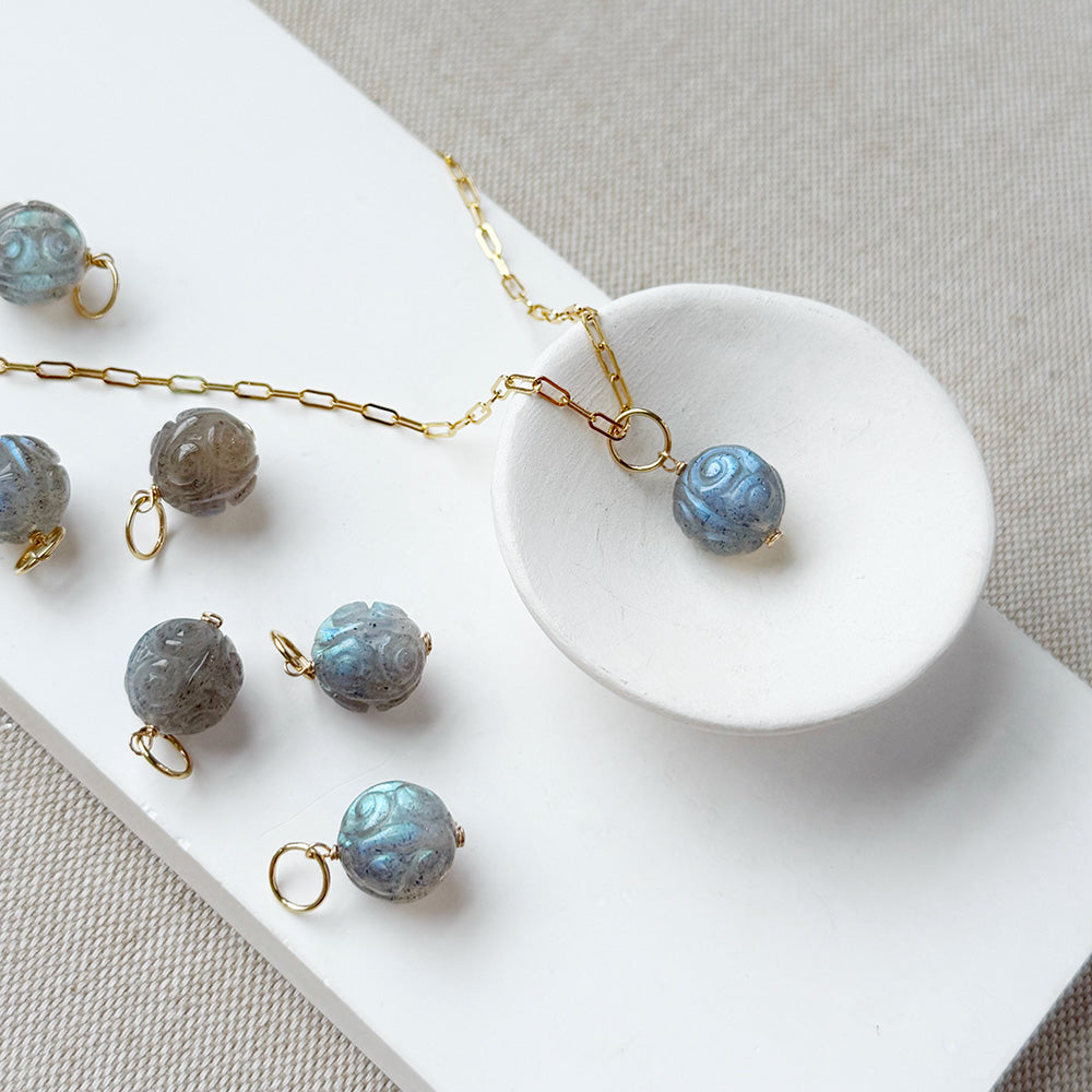 A gold chain necklace with a round carved labradorite bead pendant, displayed on a white tray with several matching loose beads and small gold rings on a neutral fabric background.