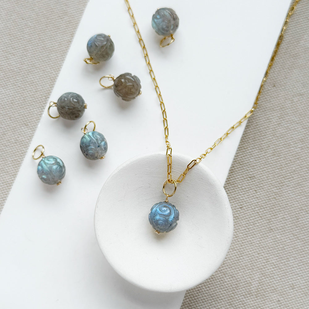A gold chain necklace with a round carved labradorite bead pendant, displayed on a white tray with several matching loose beads and small gold rings on a neutral fabric background.