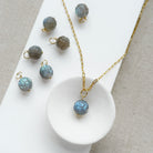 A gold chain necklace with a round carved labradorite bead pendant, displayed on a white tray with several matching loose beads and small gold rings on a neutral fabric background.