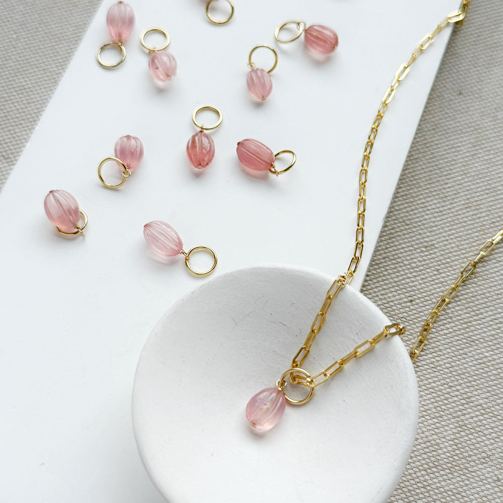 Gold chain necklace with a small faceted pink bead pendant, displayed on a white dish with several matching pink bead charms scattered on a white board.