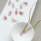 Gold chain necklace with a small faceted pink bead pendant, displayed on a white dish with several matching pink bead charms scattered on a white board.