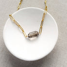 A gold paperclip chain necklace with a center set smokey quartz, displayed on a white card over a textured gray fabric background.