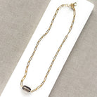 A gold paperclip chain necklace with a center set smokey quartz, displayed on a white card over a textured gray fabric background.