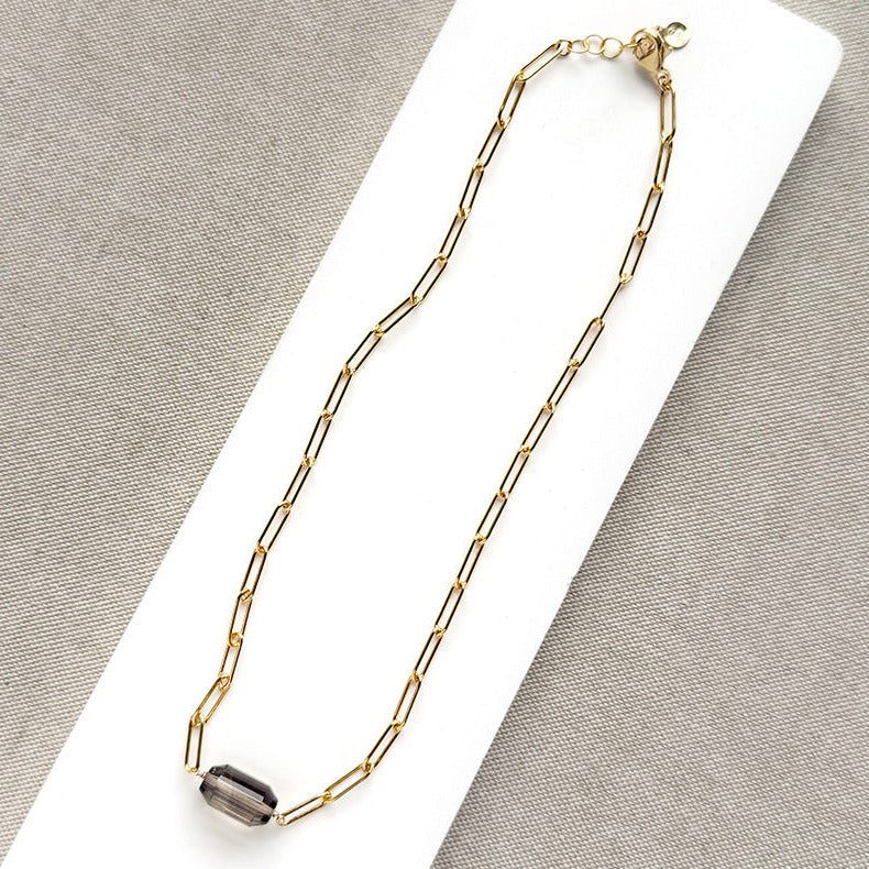 A gold paperclip chain necklace with a center set smokey quartz, displayed on a white card over a textured gray fabric background.