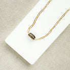 A gold paperclip chain necklace with a center set smokey quartz, displayed on a white card over a textured gray fabric background.