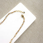 A gold paperclip chain necklace with a lobster clasp and extender, laid on a white rectangular card over a textured beige fabric background.