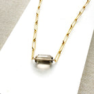 A gold paperclip chain necklace with a center set smokey quartz, displayed on a white card over a textured gray fabric background.