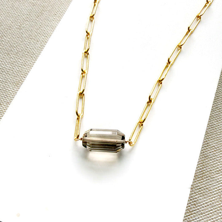 A gold paperclip chain necklace with a center set smokey quartz, displayed on a white card over a textured gray fabric background.