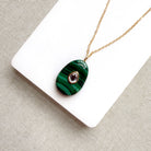 A gold chain necklace with an oval green malachite pendant featuring a small teardrop-shaped purple gemstone in the center, displayed on a white card over a textured beige fabric background.