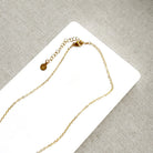 A delicate gold chain necklace with an adjustable lobster clasp and a small round engraved charm, displayed on a white card over a textured gray fabric background.