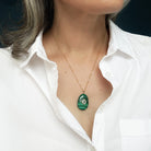 A close-up of a person wearing a delicate gold chain necklace with an oval green stone pendant featuring a small white inlay, styled with a white button-down shirt.