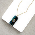 A gold chain necklace with a rectangular dark teal and green stone pendant featuring a small gold-set clear gemstone, displayed on a white card over a textured gray fabric background.