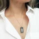 A close-up of a person wearing a delicate gold chain necklace with a rectangular dark green pendant featuring a small clear stone in the center, styled with a white collared shirt.