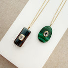 Two gold pendant necklaces displayed on a white card over a beige woven fabric background. One necklace has a dark green and black striped rectangular pendant with a small clear stone.