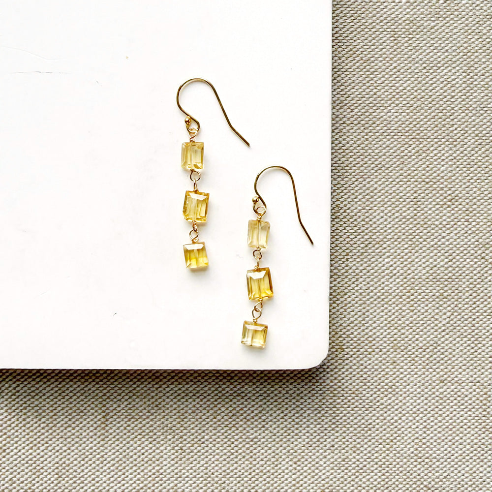 A pair of gold-tone dangle earrings with three small rectangular citrine gemstones each, displayed on a white rectangular surface over a textured beige fabric background.