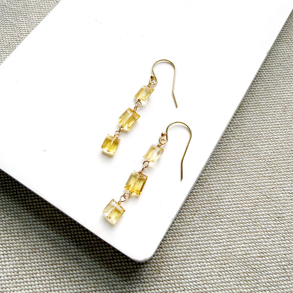 A pair of gold-tone dangle earrings with three small rectangular citrine gemstones each, displayed on a white rectangular surface over a textured beige fabric background.