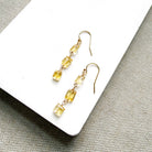 A pair of gold-tone dangle earrings with three small rectangular citrine gemstones each, displayed on a white rectangular surface over a textured beige fabric background.