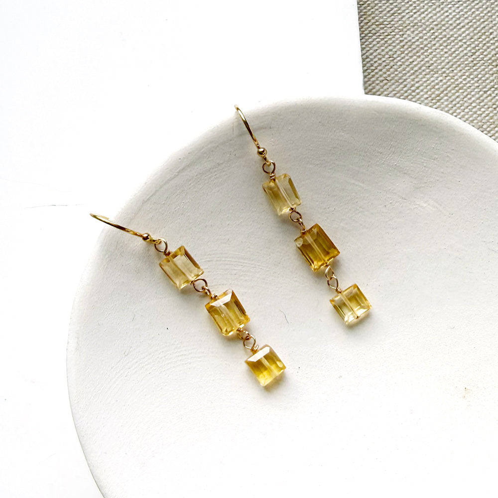 A pair of gold-tone dangle earrings with three small rectangular citrine gemstones each, displayed on a white rectangular surface over a textured beige fabric background.