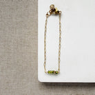Gold chain bracelet with green beads on a white card against a beige fabric background