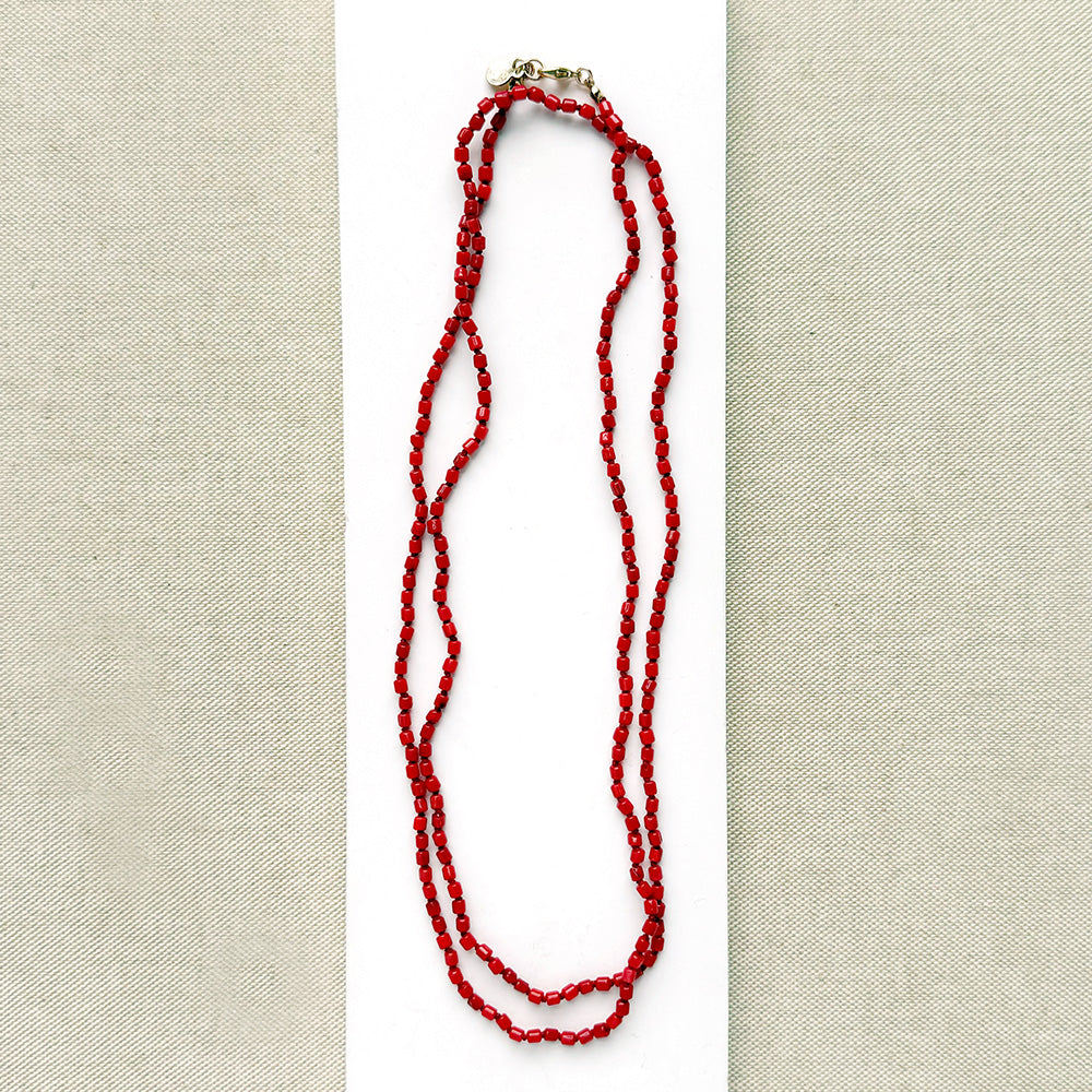 A long knotted necklace made of small red coral beads placed on a white rectangular display card over a textured light gray fabric background.
