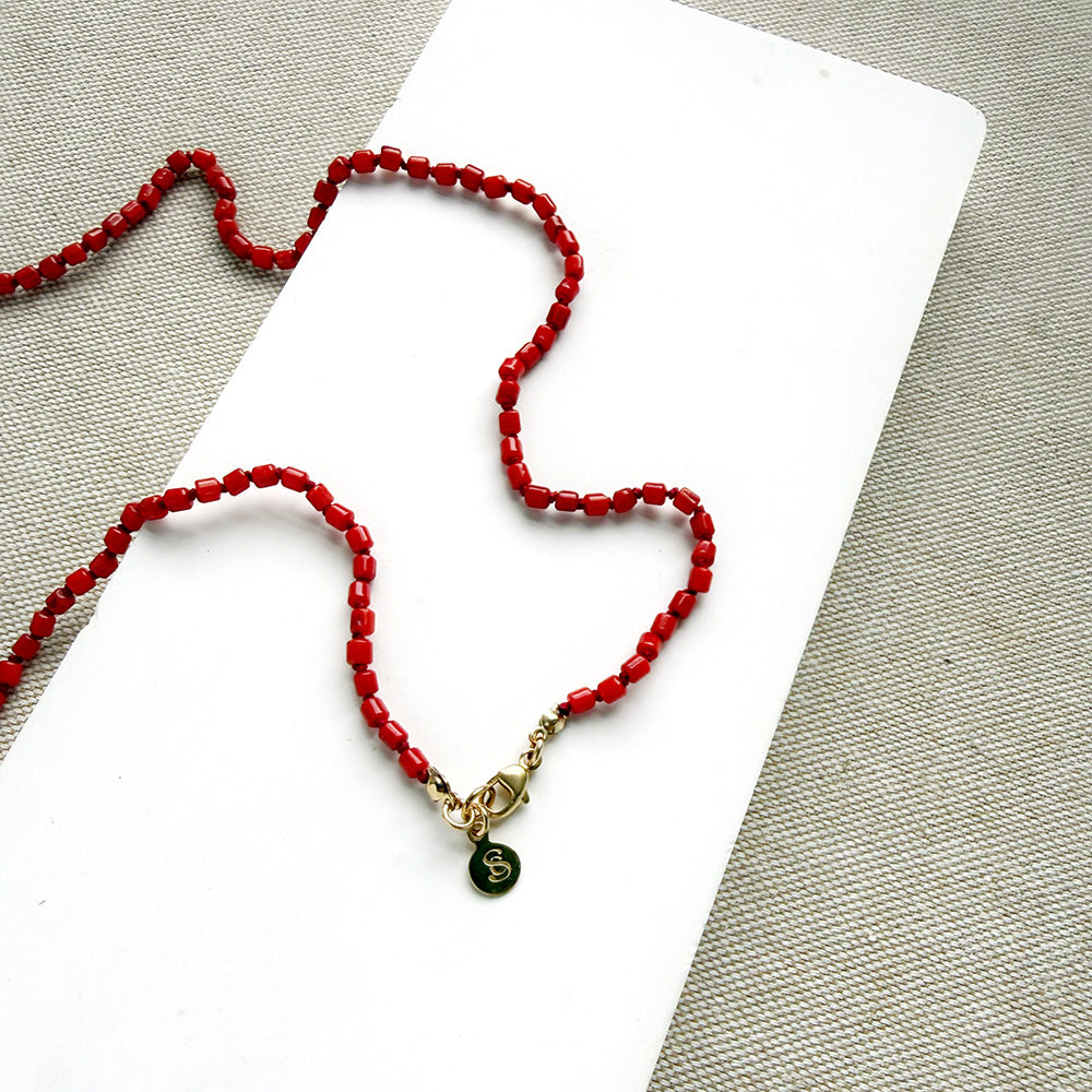A long knotted necklace made of small red coral beads placed on a white rectangular display card over a textured light gray fabric background.