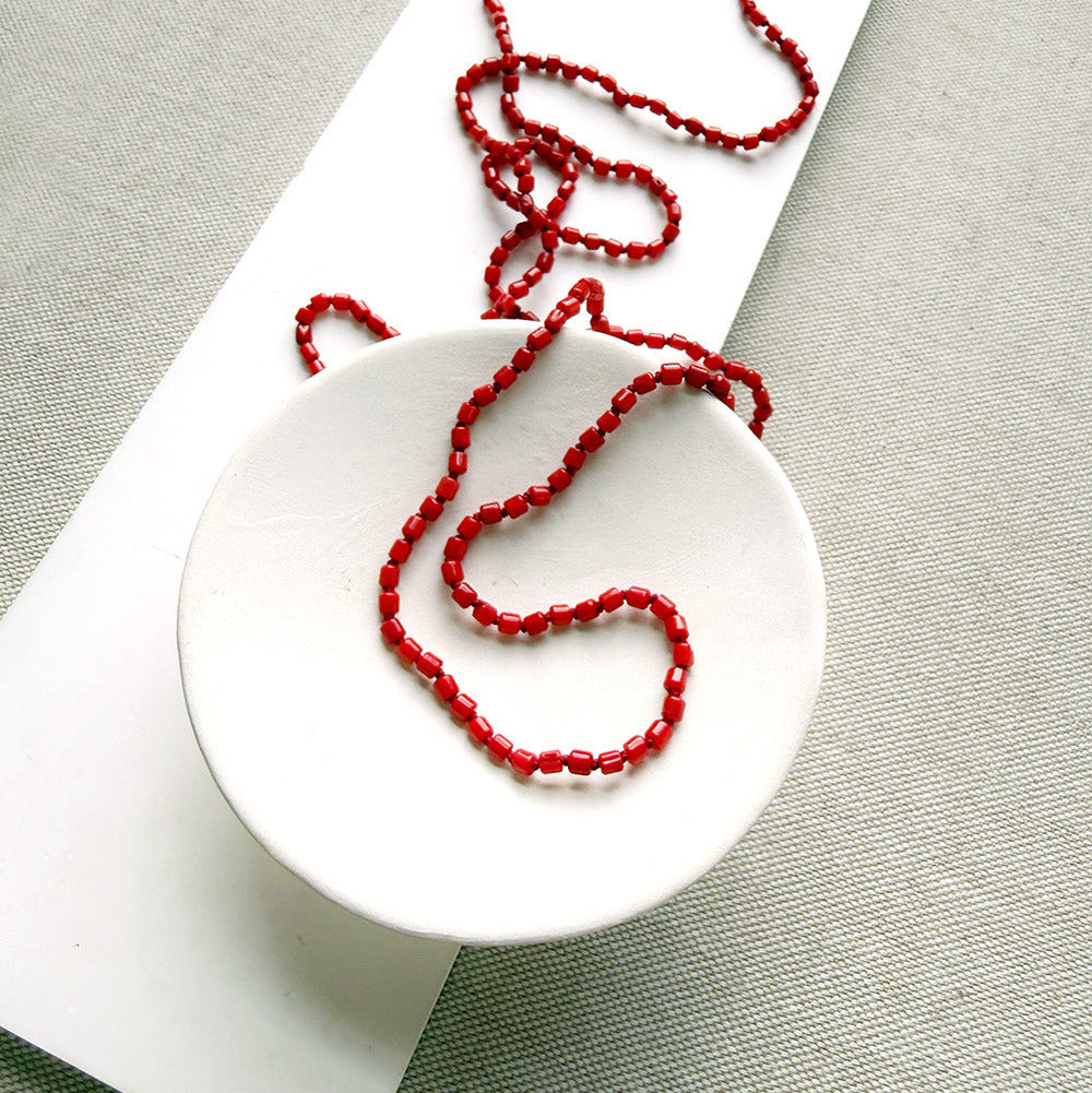 A long knotted necklace made of small red coral beads draped over a small white ceramic dish, placed on a white rectangular display card over a textured light gray fabric background.