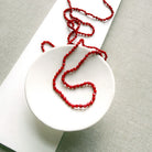 A long knotted necklace made of small red coral beads draped over a small white ceramic dish, placed on a white rectangular display card over a textured light gray fabric background.