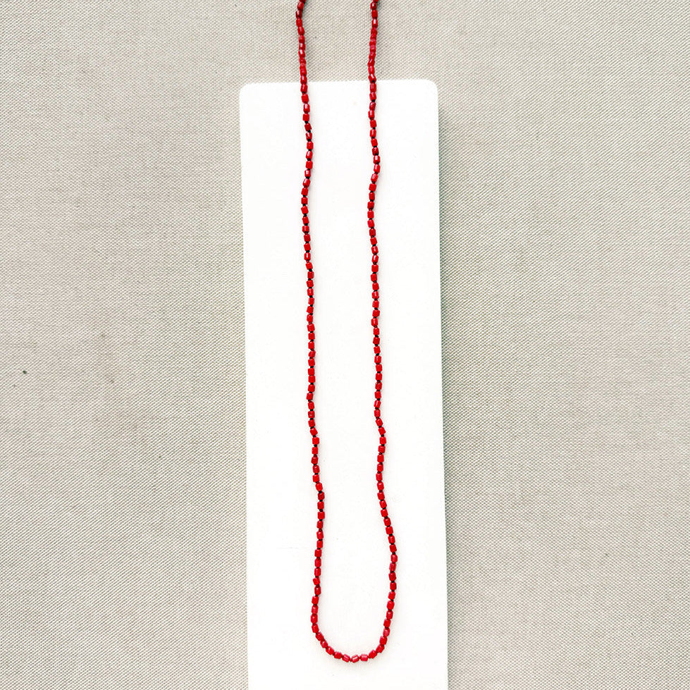 A long knotted necklace made of small red coral beads placed on a white rectangular display card over a textured light gray fabric background.