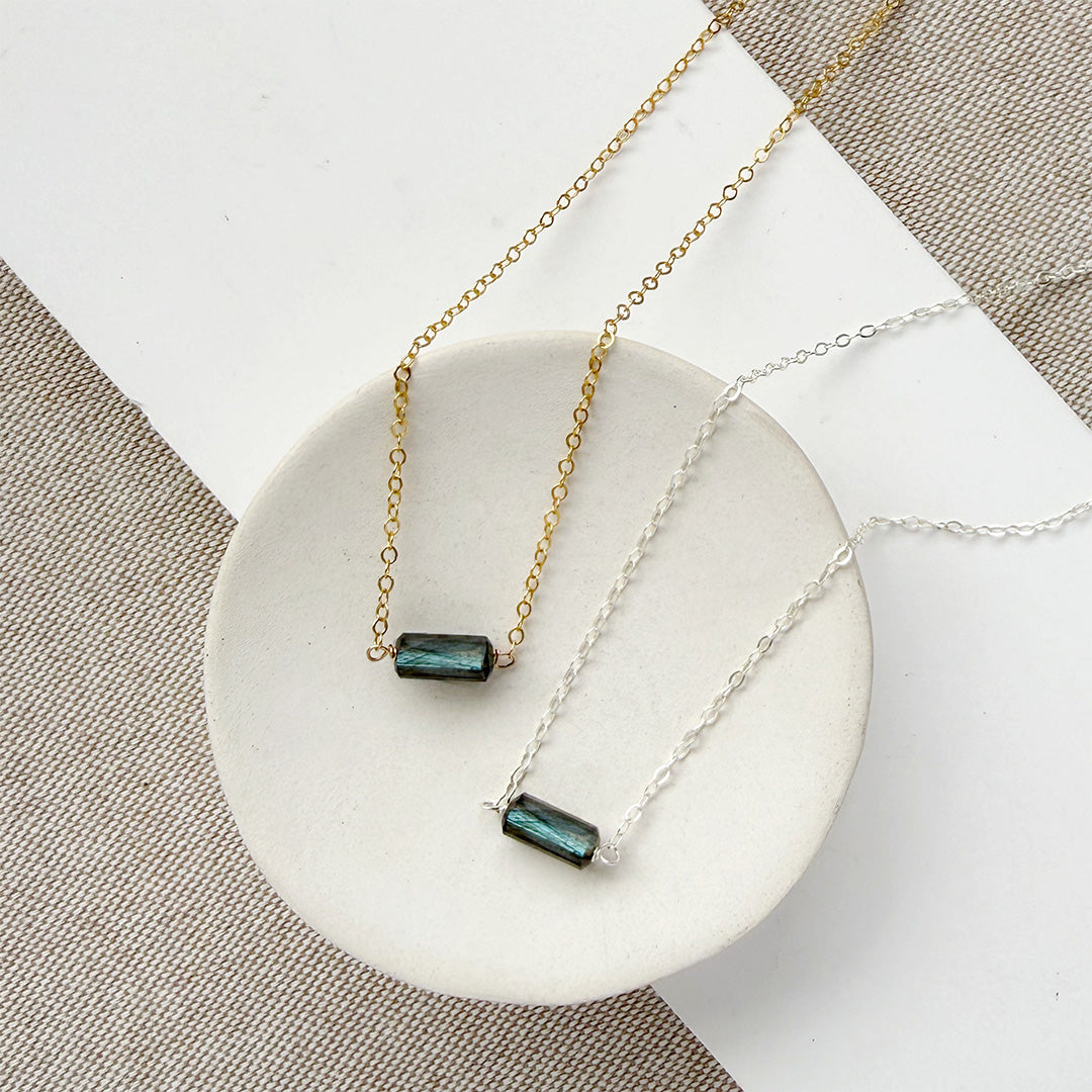 Two necklaces, one silver and one gold, with a single labradorite gemstone on a white dish against a textured surface