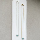 Two necklaces, one silver and one gold, with a single labradorite gemstone on a white dish against a textured surface