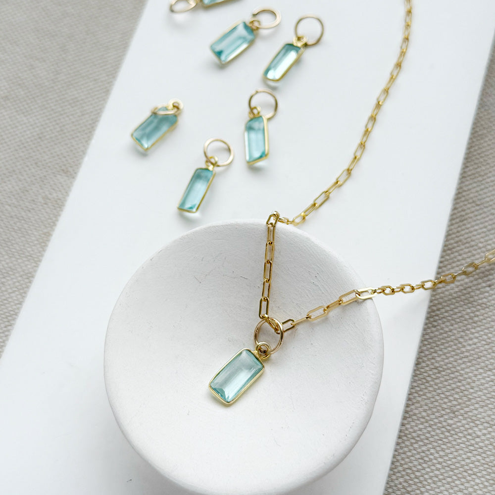 Gold-tone necklace with emerald cut aquamarine charm, displayed on a white dish and matching white rectangular board over a neutral fabric background.