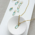 Gold-tone necklace with emerald cut aquamarine charm, displayed on a white dish and matching white rectangular board over a neutral fabric background.