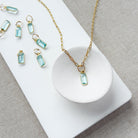 Gold-tone necklace with emerald cut aquamarine charm, displayed on a white dish and matching white rectangular board over a neutral fabric background.