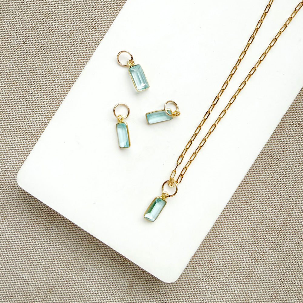 Gold-tone necklace with emerald cut aquamarine charm, displayed on a white rectangular board over a neutral fabric background.