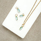 Gold-tone necklace with emerald cut aquamarine charm, displayed on a white rectangular board over a neutral fabric background.