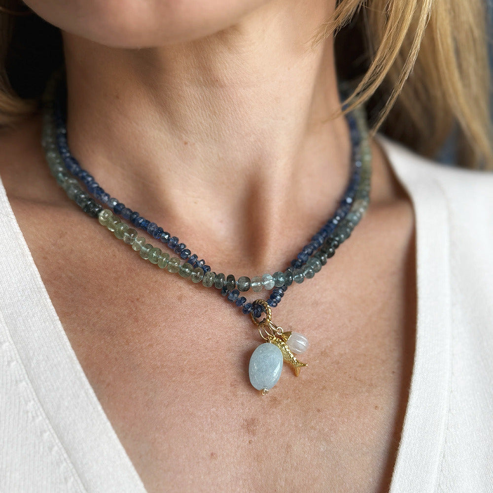 A close-up of a woman wearing a multi-strand beaded necklace with a light blue oval pendant and gold-toned accents, paired with a white V-neck top.