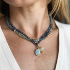 A close-up of a woman wearing a multi-strand beaded necklace with a light blue oval pendant and gold-toned accents, paired with a white V-neck top.